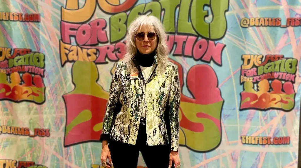 Marlene Weisman wearing circular sunglasses and a patterned, light-colored jacket stands before a bright “The Fest for Beatles Fans Revolution 2025” step‑and‑repeat banner with bubble lettering and Beatles Fest logos