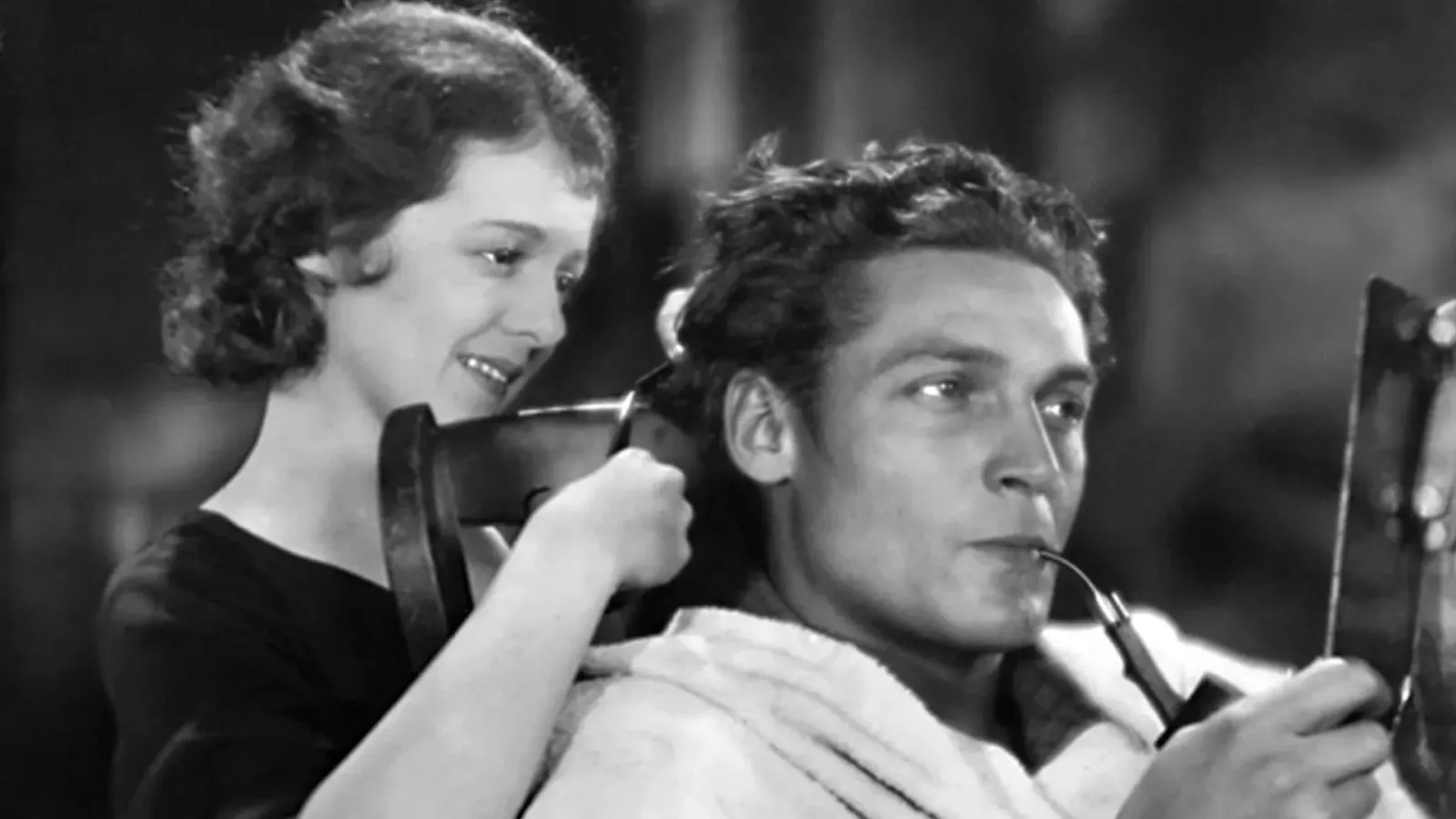 Black‑and‑white still of Janet Gaynor smiling as she trims Charles Farrell’s hair while he smokes a pipe and looks into a small mirror, from 7th (1927) Heaven directed by Frank Borzage.