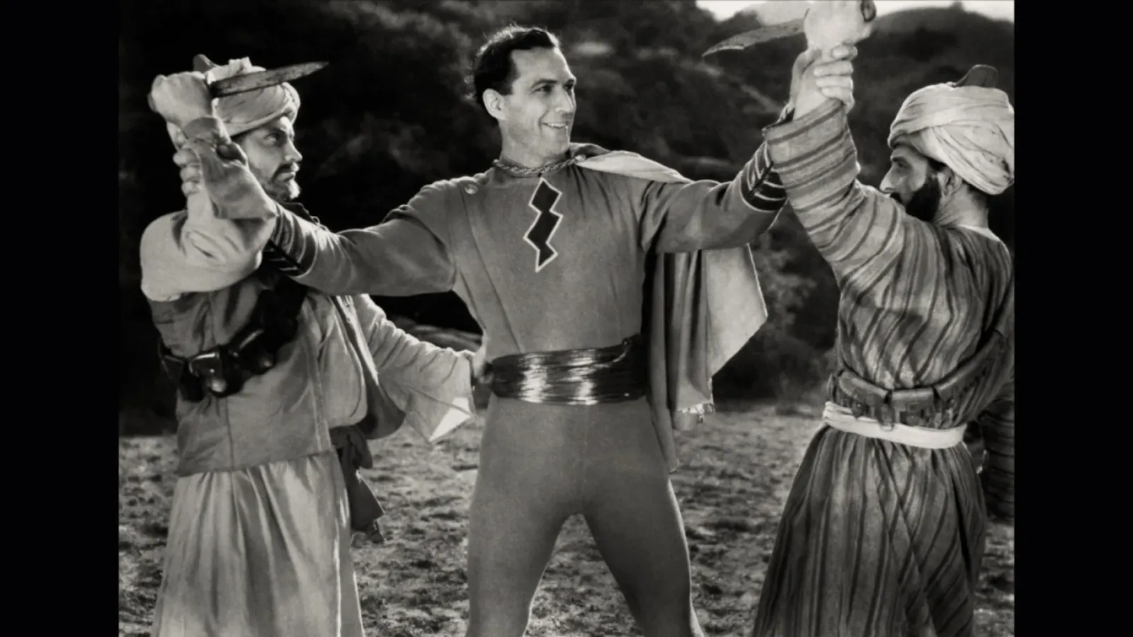 Tom Tyler in the Captain Marvel costume, arms outstretched as he stops two knife‑wielding attackers, from The Adventures of Captain Marvel.